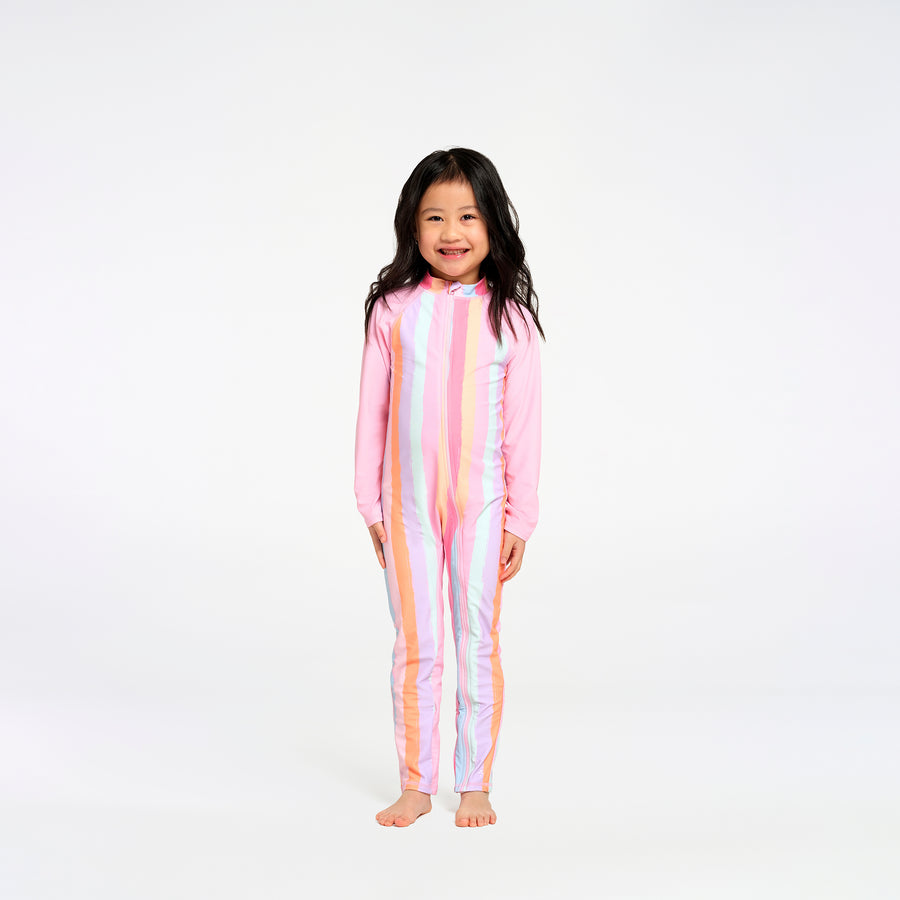 A young girl with long dark hair stands barefoot against a plain white studio background, wearing a one-piece long-sleeve swimsuit with vertical pastel stripes