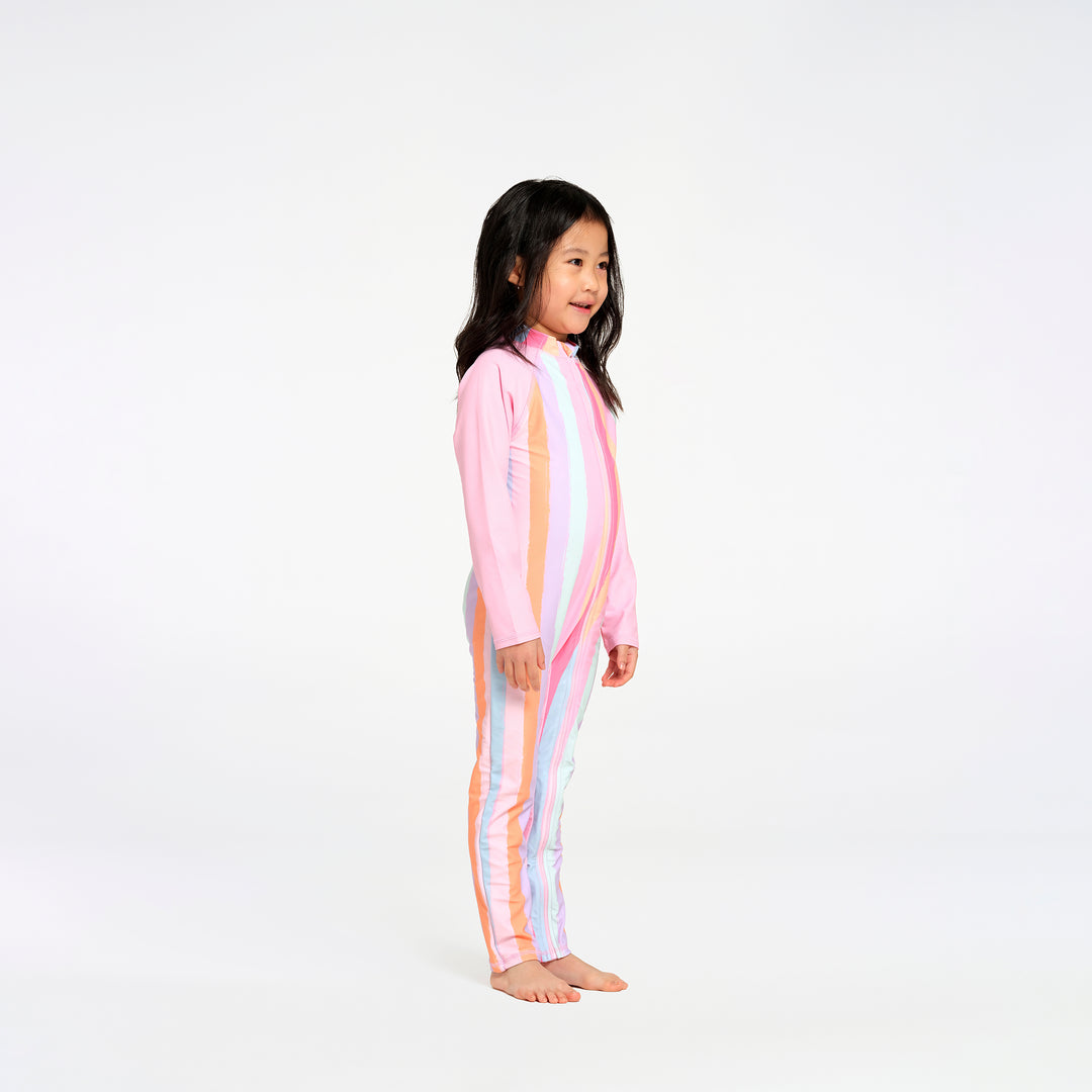 A young girl stands in profile wearing a one-piece long-sleeve swim suit with pastel vertical stripes in pink, orange, yellow, and blue on a solid white studio background.