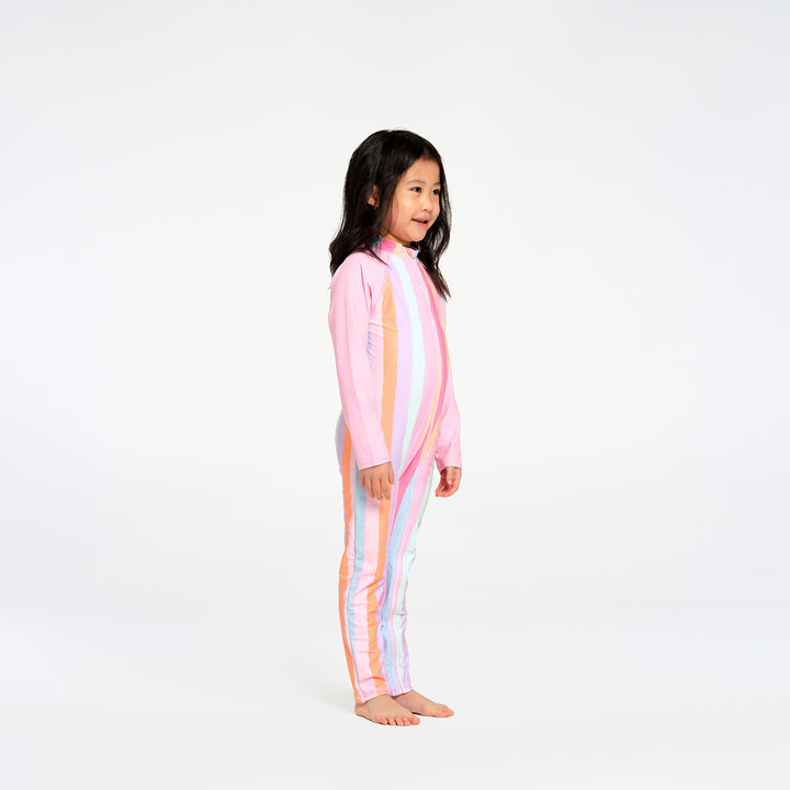 A young girl stands in profile wearing a one-piece long-sleeve swim suit with pastel vertical stripes in pink, orange, yellow, and blue on a solid white studio background.