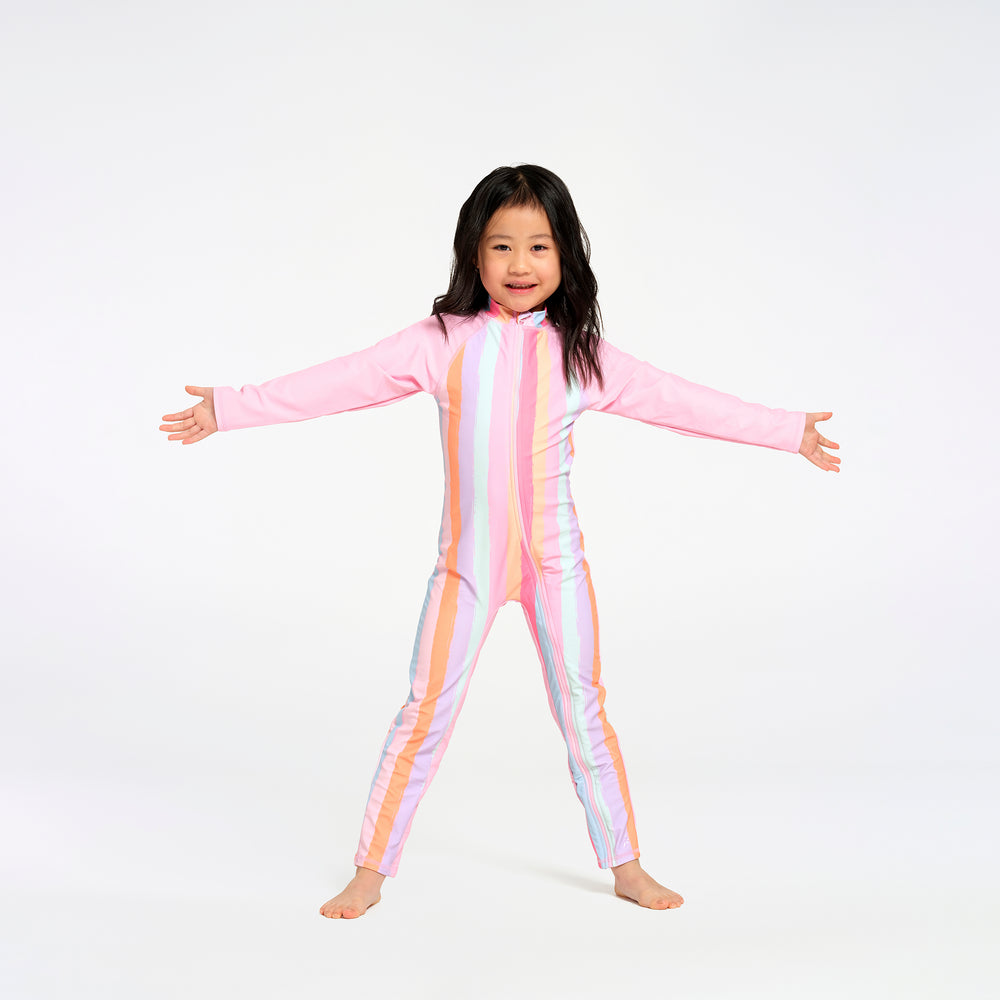 A young girl with dark hair stands barefoot against a white studio background, wearing a full-body long-sleeve one-piece swimsuit with vertical pastel stripes in pink, orange, yellow, and white.