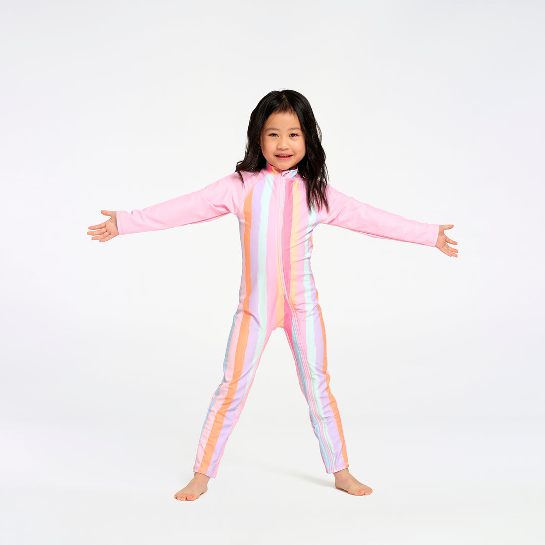 A young girl with dark hair stands barefoot against a white studio background, wearing a full-body long-sleeve one-piece swimsuit with vertical pastel stripes in pink, orange, yellow, and white.