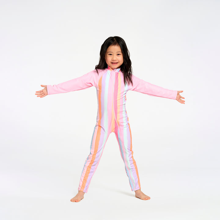 A young girl with dark hair stands barefoot against a white studio background, wearing a full-body long-sleeve one-piece swimsuit with vertical pastel stripes in pink, orange, yellow, and white.
