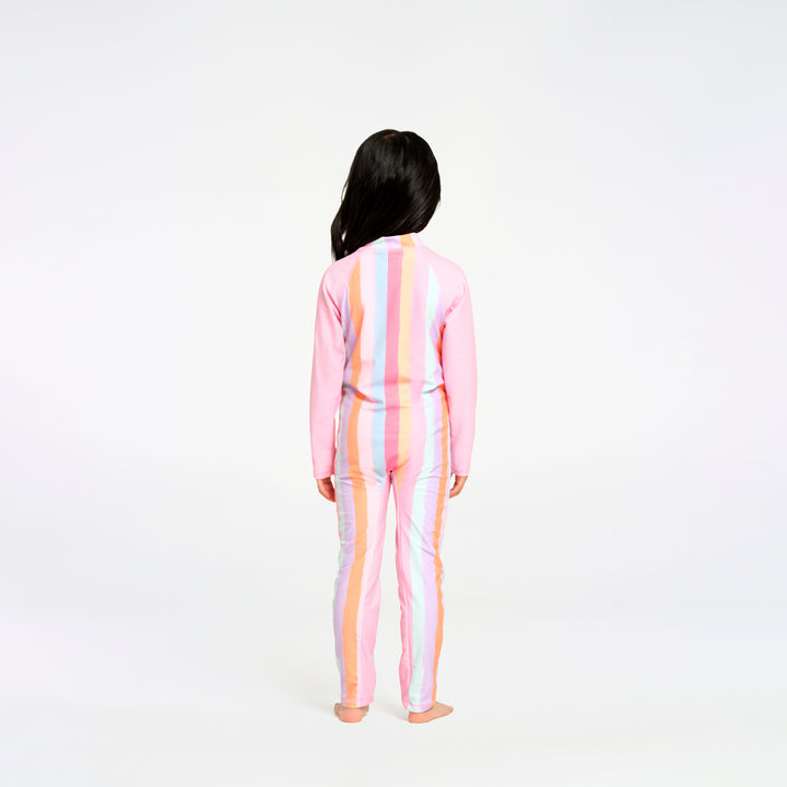 A child standing with their back to the camera, wearing a pink long-sleeve one-piece swimsuit with vertical pastel stripes in blue, yellow, and orange on a plain white studio background.