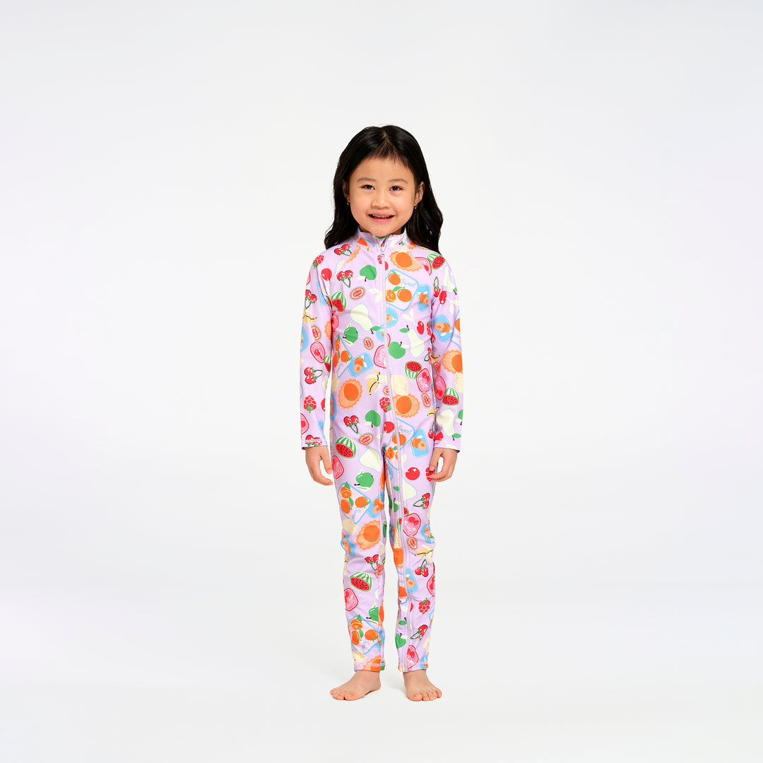 A young girl with long black hair stands barefoot against a plain white studio background, wearing a one-piece long-sleeve swimsuit with a colorful fruit print on a light pink base.