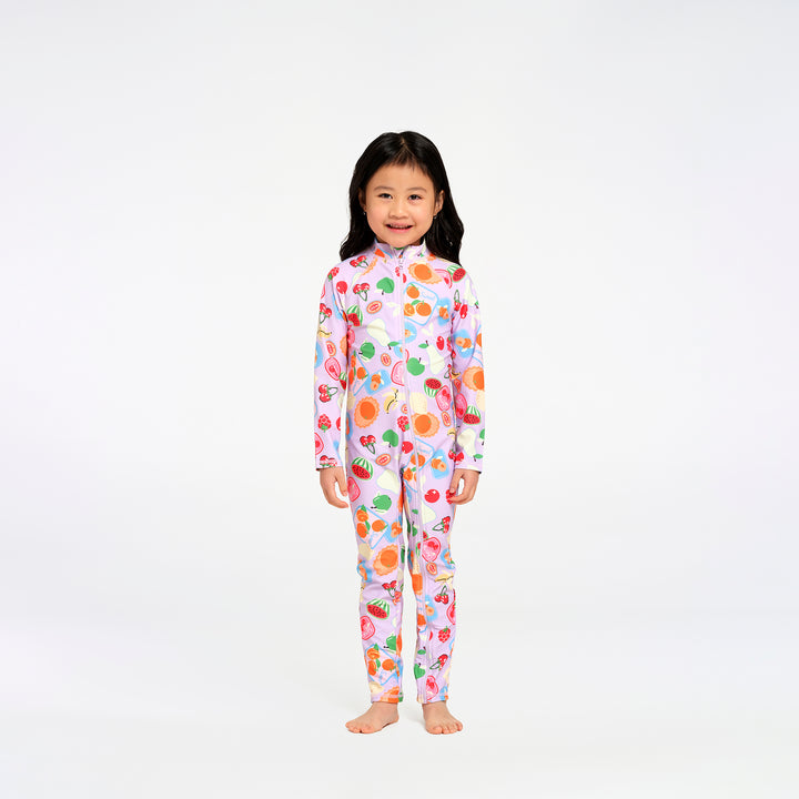 A young girl with long black hair stands barefoot against a plain white studio background, wearing a one-piece long-sleeve swimsuit with a colorful fruit print on a light pink base.