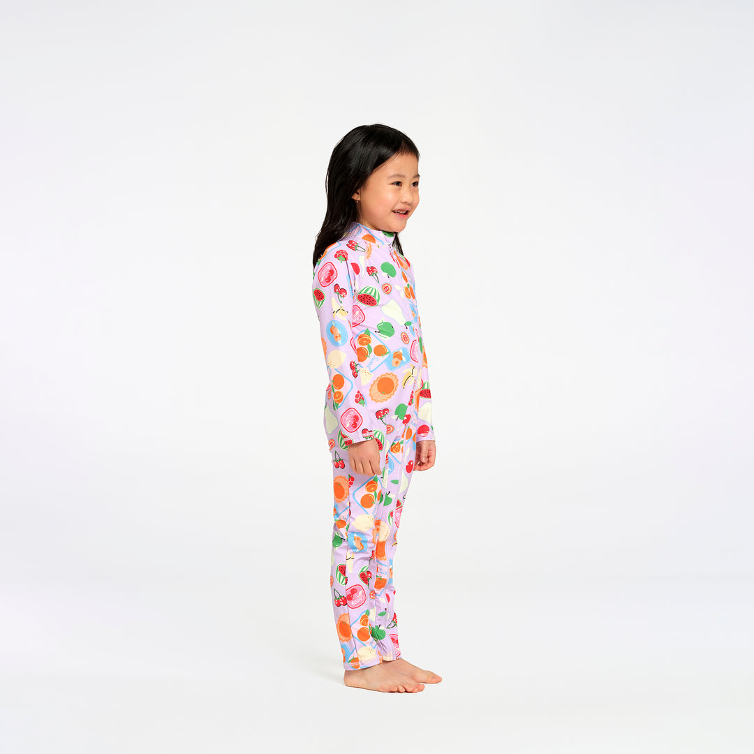 A young girl with long black hair stands barefoot against a plain white studio background, wearing a one-piece long-sleeve swimsuit with a colorful fruit print on a light pink base.