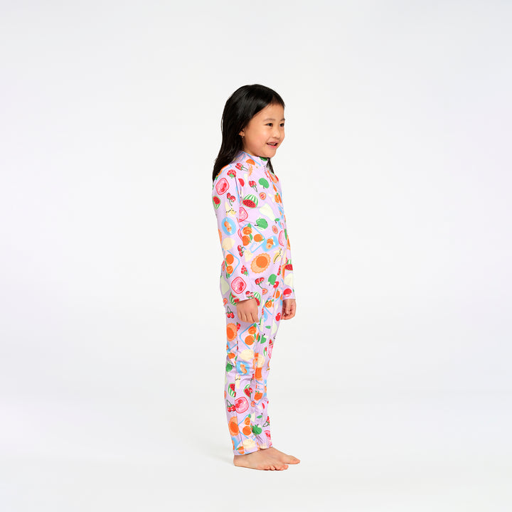 A young girl with long black hair stands barefoot against a plain white studio background, wearing a one-piece long-sleeve swimsuit with a colorful fruit print on a light pink base.