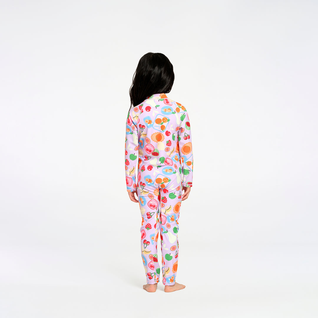 A child standing with their back to the camera, wearing a matching long-sleeve swimsuit with a colorful fruit and dessert print on a light pink background, set against a plain white studio backdrop.