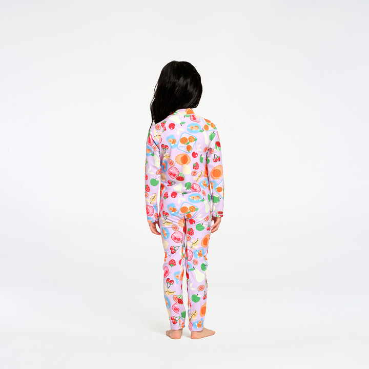 A child standing with their back to the camera, wearing a matching long-sleeve swimsuit with a colorful fruit and dessert print on a light pink background, set against a plain white studio backdrop.