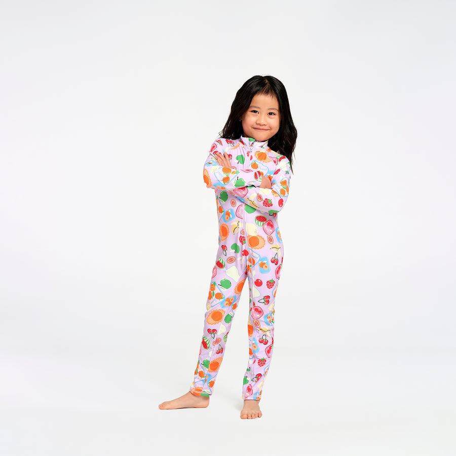A young girl with long black hair stands barefoot against a plain white studio background, wearing a one-piece long-sleeve swimsuit with a colorful fruit print on a light pink base.