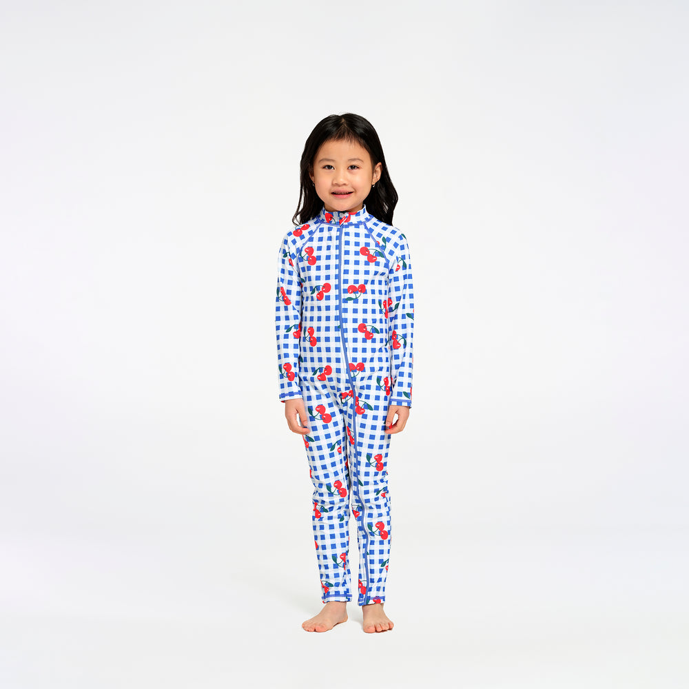 A young girl stands barefoot against a plain white studio background, wearing a one-piece long-sleeve swimsuit with a blue gingham pattern and scattered red cherry motifs.