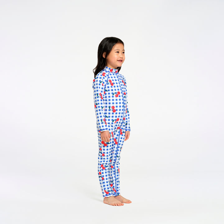 A young girl stands in profile wearing a long-sleeve swimsuit with a blue and white gingham pattern and small red cherry motifs, photographed against a seamless white studio background.