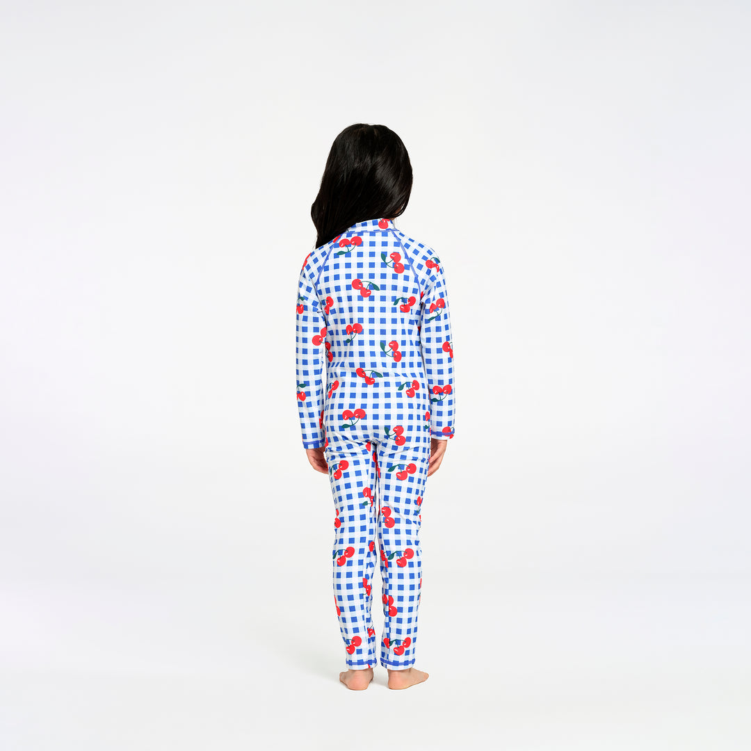 A child standing with their back to the camera, wearing a matching long-sleeve swimsuit with a blue and white gingham pattern and red cherry motifs, set against a plain white studio background.