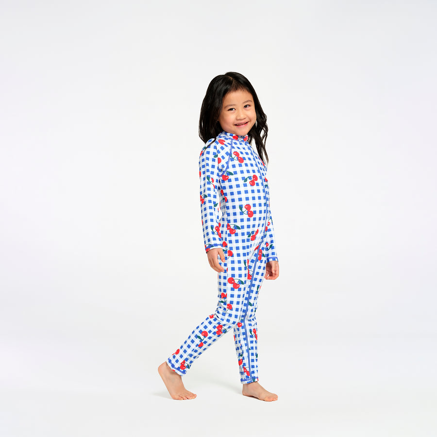 A young girl with dark hair stands barefoot in a studio setting, wearing a one-piece long-sleeve swimsuit with a blue and white gingham pattern and red cherry motifs.
