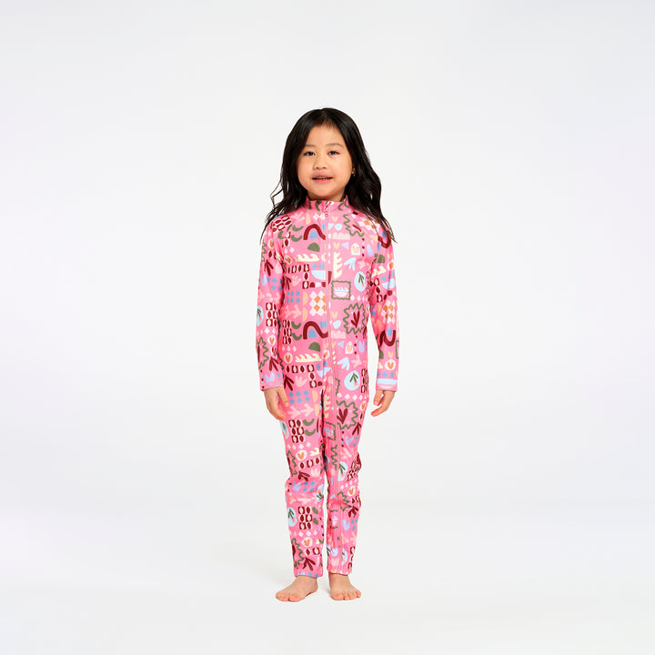 A young girl stands barefoot against a plain white studio background, wearing a pink long-sleeve one-piece swimsuit with a colorful abstract print.