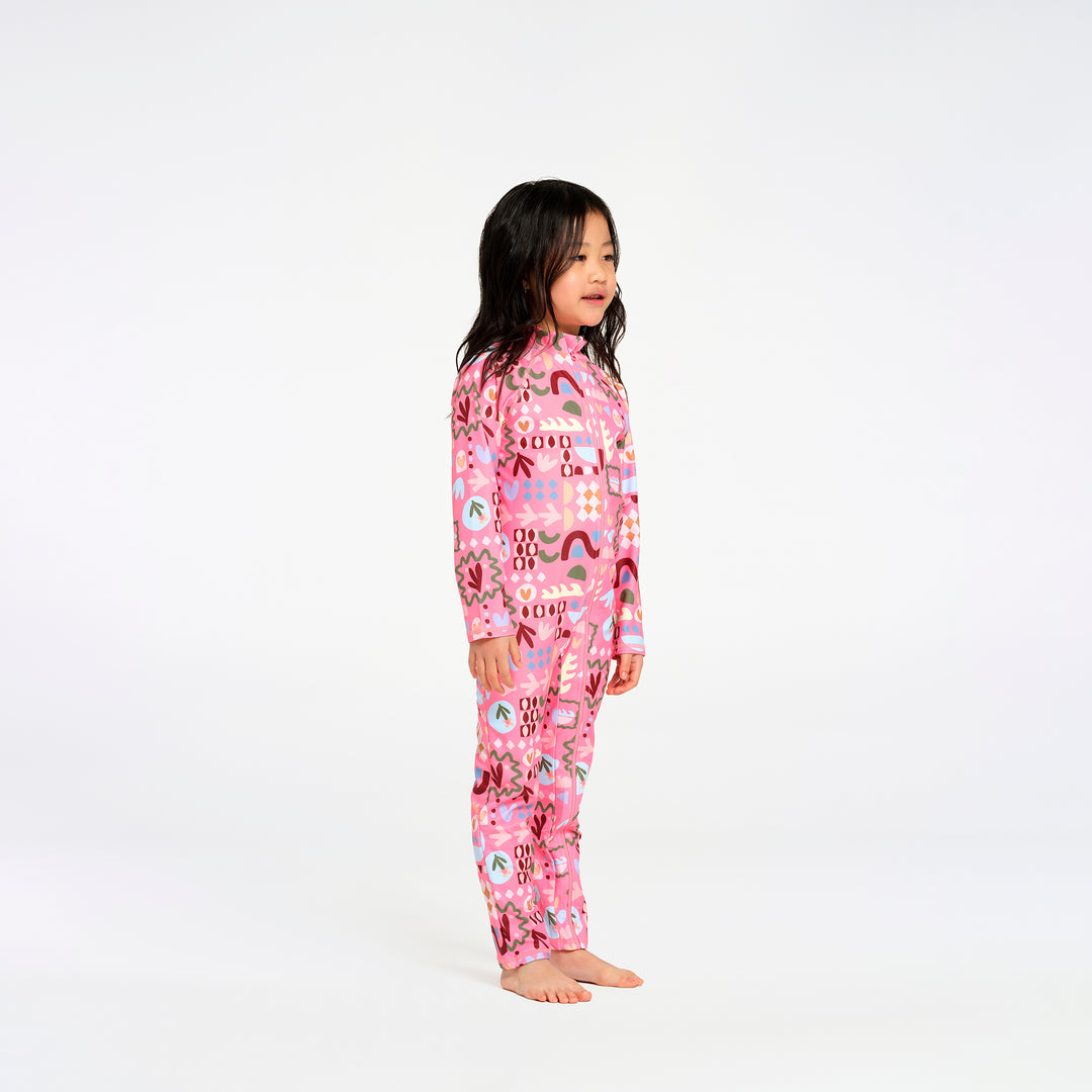 A young girl stands in profile wearing a pink long-sleeve swimsuit with a colorful rainbow pattern, photographed against a seamless white studio background.