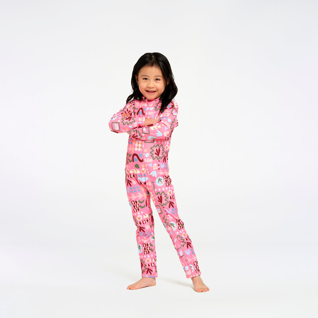 A young girl with long black hair stands barefoot against a plain white studio background, wearing a pink swimsuit with a colorful abstract print.
