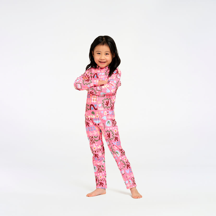 A young girl with long black hair stands barefoot against a plain white studio background, wearing a pink swimsuit with a colorful abstract print.
