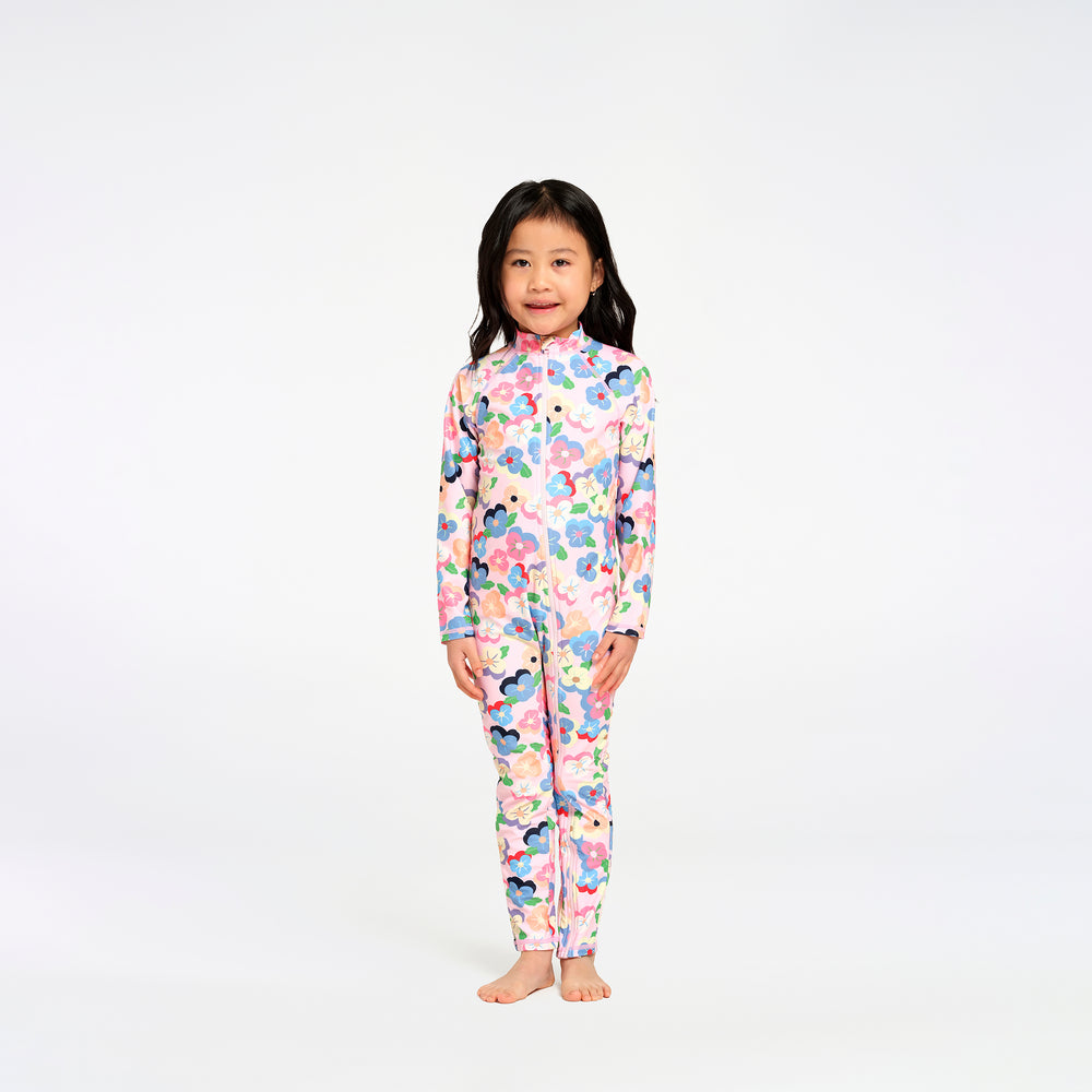 A young girl with long black hair stands barefoot against a plain white studio background, wearing a long-sleeve, high-neck one-piece swimsuitwith a colourful floral print on a light pink base.