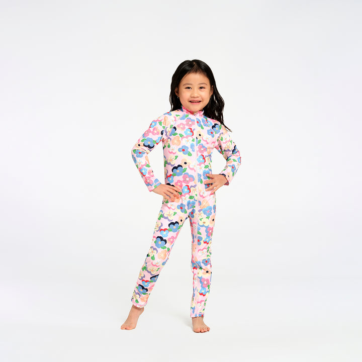 A young girl with long black hair stands barefoot against a plain white studio background, wearing a long-sleeve, high-neck one-piece swimsuitwith a colourful floral print on a light pink base.