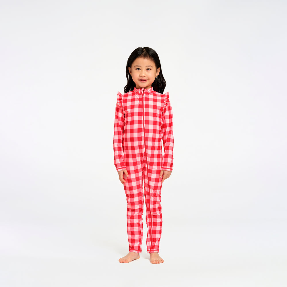 A young girl stands barefoot against a plain white studio background, wearing a red and white gingham one-piece swimsuit with long sleeves and a high collar.