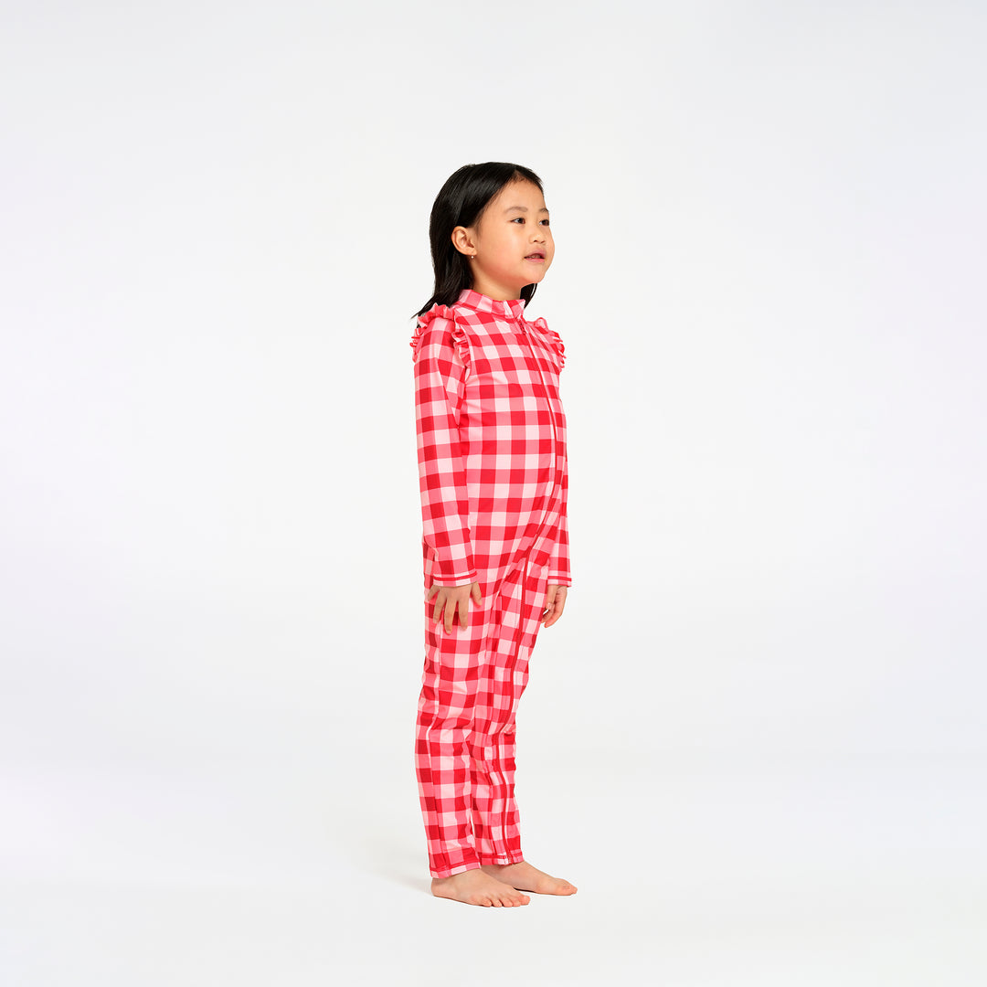 A young girl stands in profile against a seamless white studio background, wearing a red and white gingham check long-sleeve swimsuit with ruffled trim at the collar and cuffs.