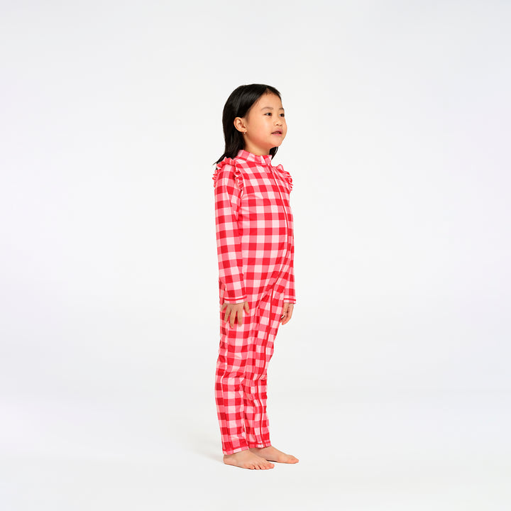 A young girl stands in profile against a seamless white studio background, wearing a red and white gingham check long-sleeve swimsuit with ruffled trim at the collar and cuffs.