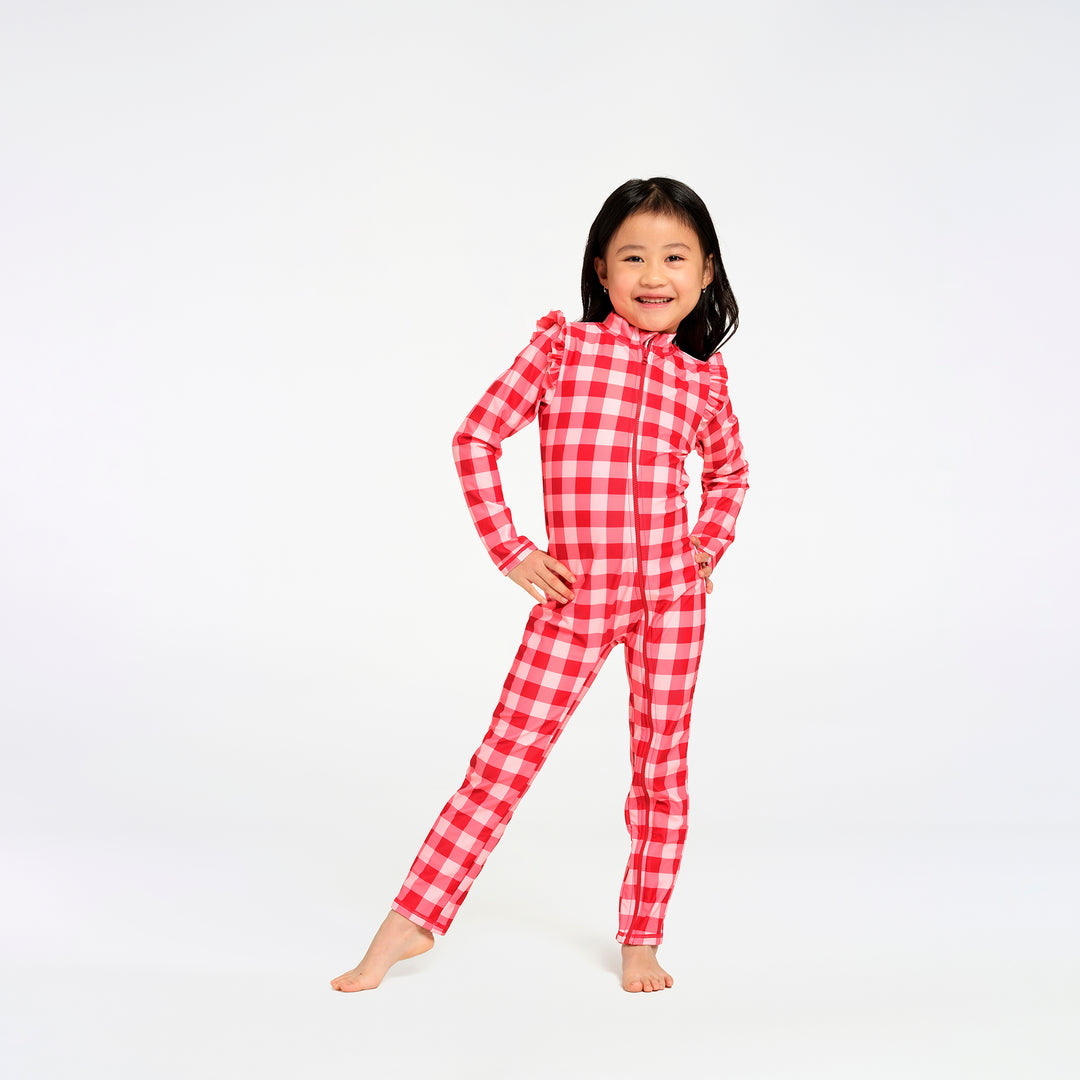A young girl stands barefoot against a plain white studio background, wearing a one-piece zip-up swimsuit in a red and white gingham check pattern.