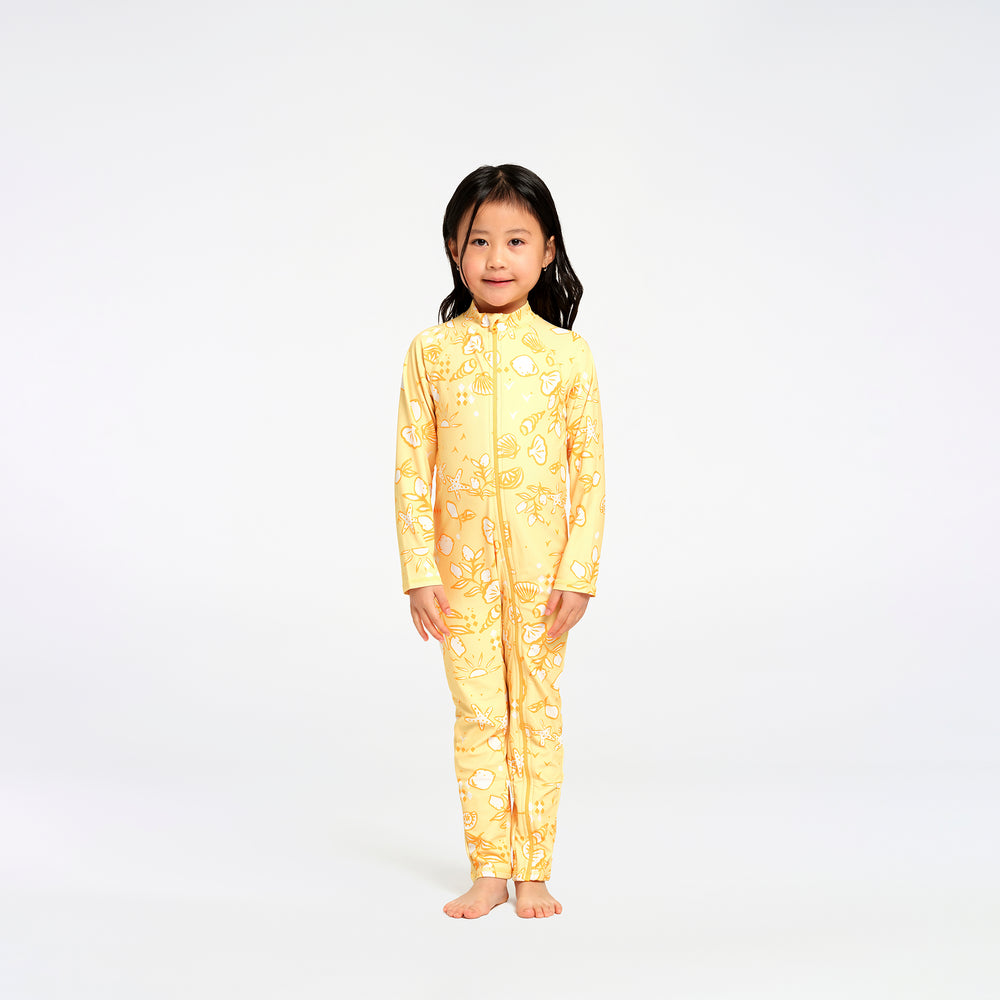 A young girl stands barefoot against a plain white studio background, wearing a full-length yellow zip-front one-piece swimsuit with an allover beach themed print.