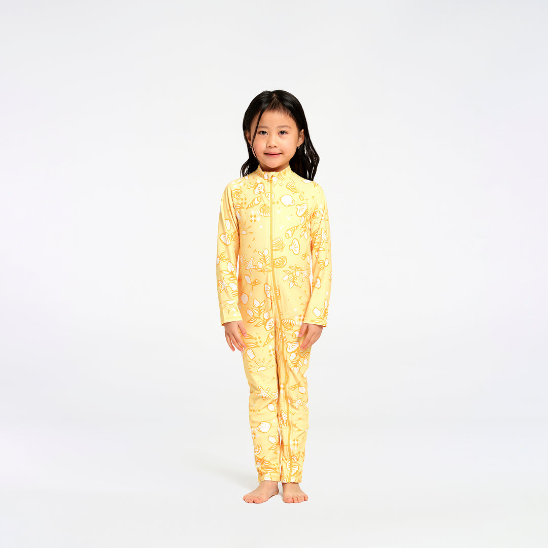 A young girl stands barefoot against a plain white studio background, wearing a full-length yellow zip-front one-piece swimsuit with an allover beach themed print.