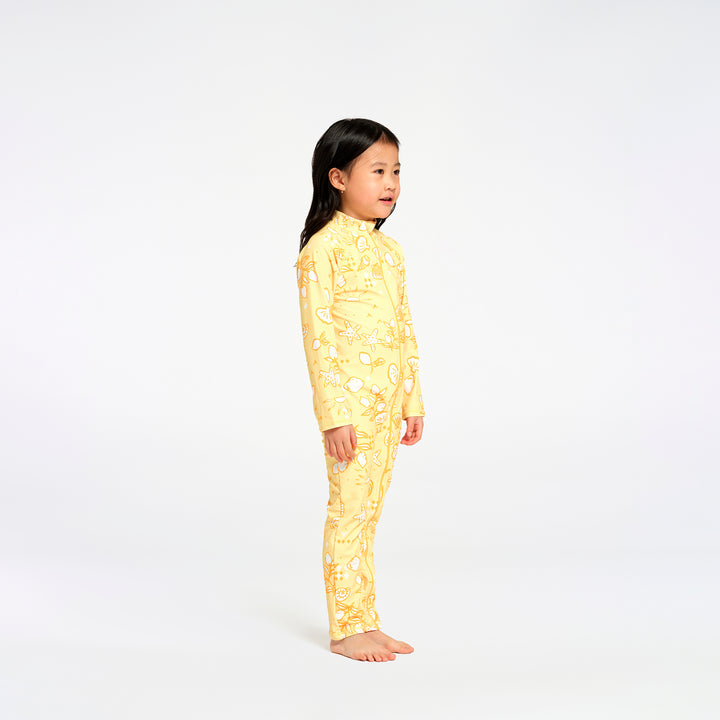 A young girl stands in profile wearing a yellow long-sleeve pajama set with a white paisley pattern against a plain white studio background.