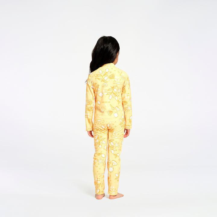 A child standing with their back to the camera, wearing a yellow long-sleeve swimsuit with a white citrus fruit pattern, set against a plain white studio background.