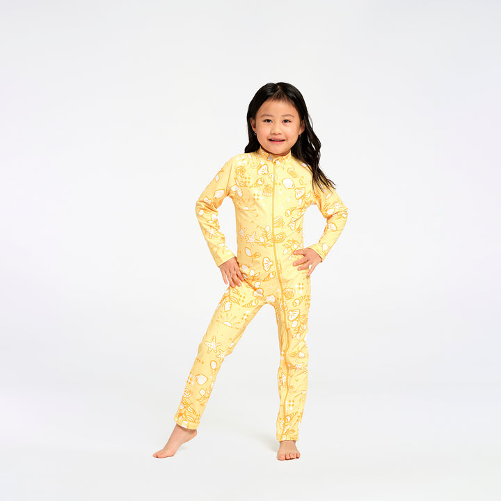 A young girl stands barefoot against a plain white studio background, wearing a full-length yellow one-piece swimsuit with an allover beach themed print.
