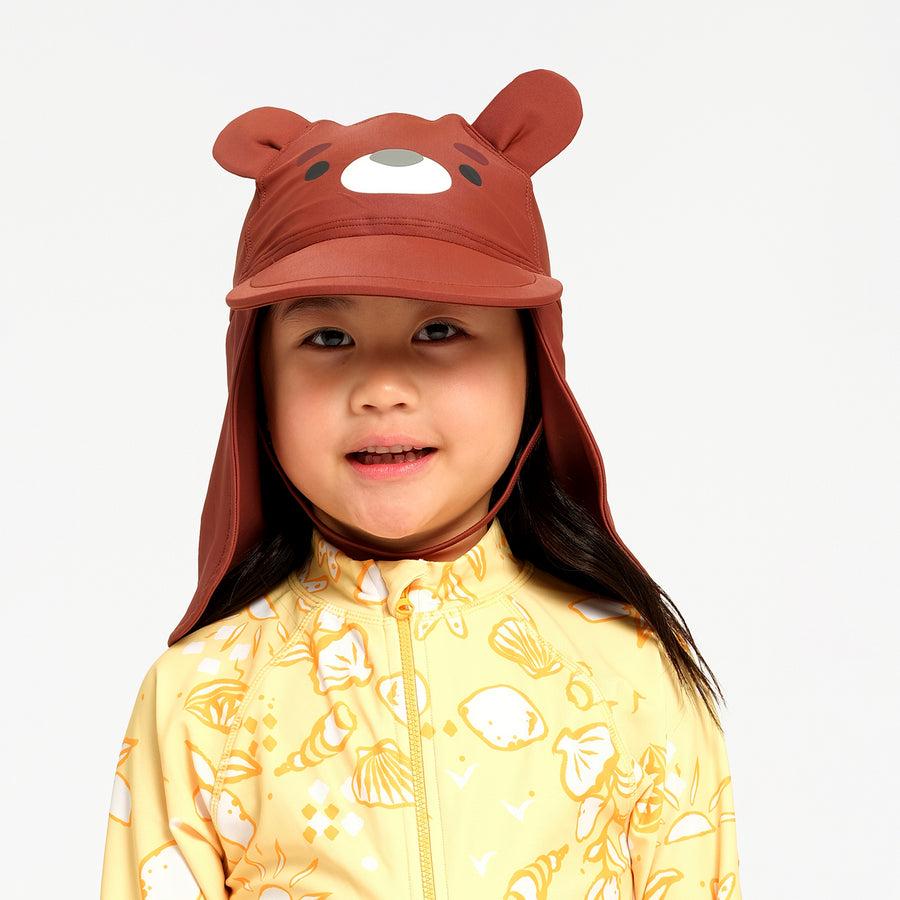 A young child wearing a brown bear-themed sun hat with a chin strap and a yellow patterned swimsuit, photographed against a clean white background.