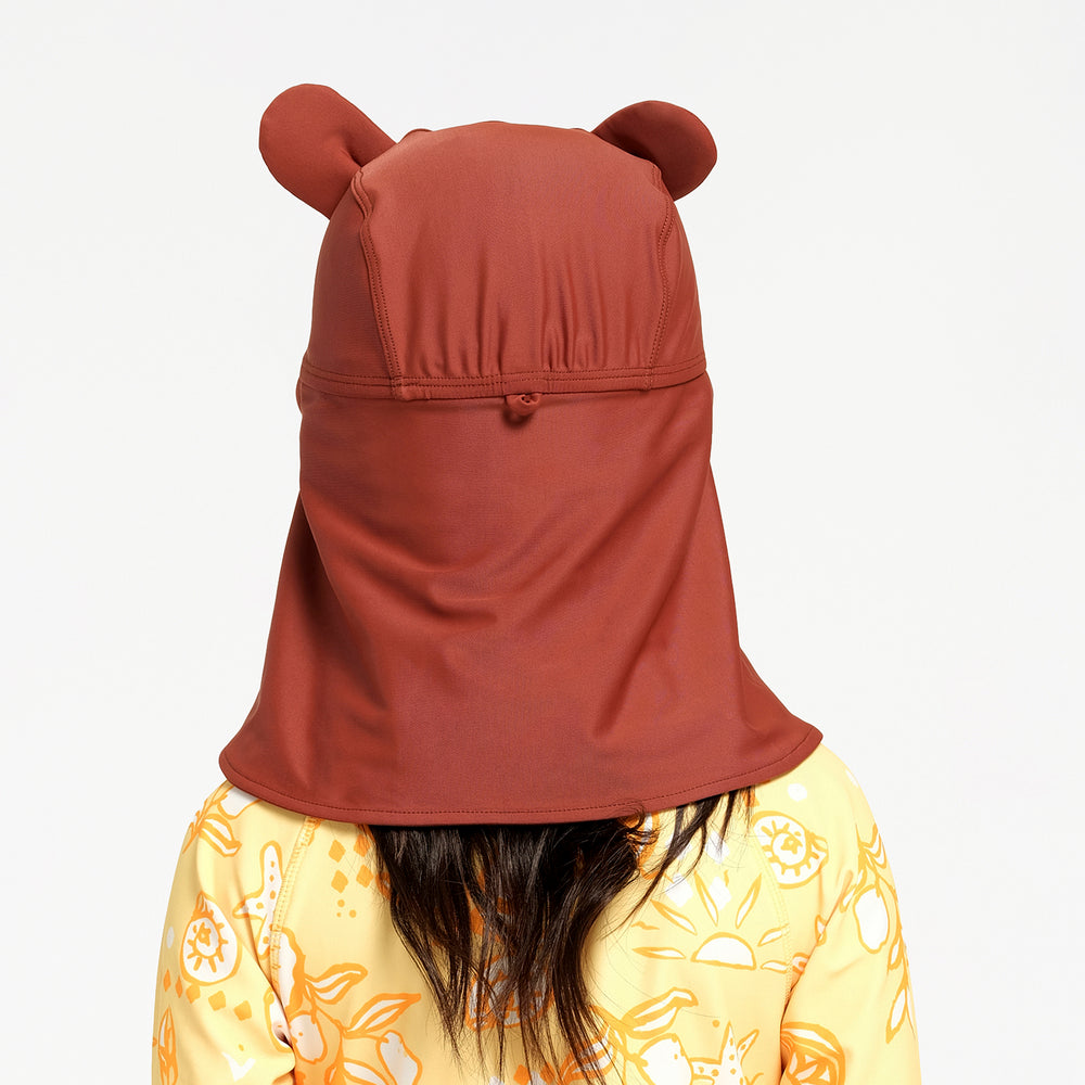 A child viewed from behind wearing a rust-brown sun hat with ear-like side protrusions and a large neck flap, over a yellow patterned shirt against a plain light gray background.