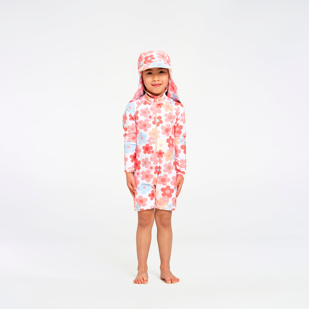 A young child standing barefoot against a plain white studio background, wearing a long-sleeve, short-legged swim suit with a matching sun hat. The swimwear has a white base with a pink and red floral pattern and small light blue accents.