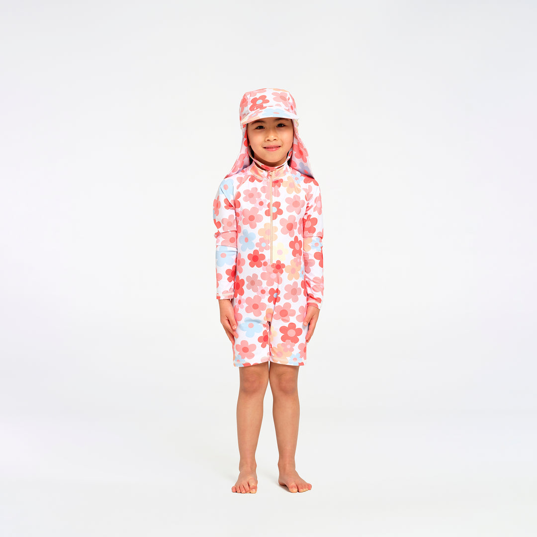 A young child standing barefoot against a plain white studio background, wearing a long-sleeve, short-legged swim suit with a matching sun hat. The swimwear has a white base with a pink and red floral pattern and small light blue accents.