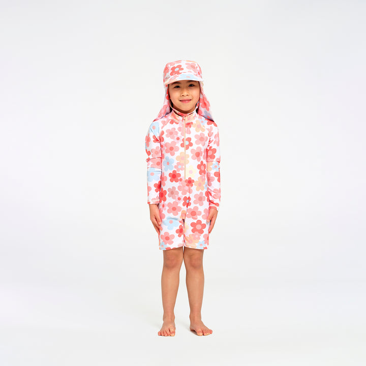 A young child standing barefoot against a plain white studio background, wearing a long-sleeve, short-legged swim suit with a matching sun hat. The swimwear has a white base with a pink and red floral pattern and small light blue accents.