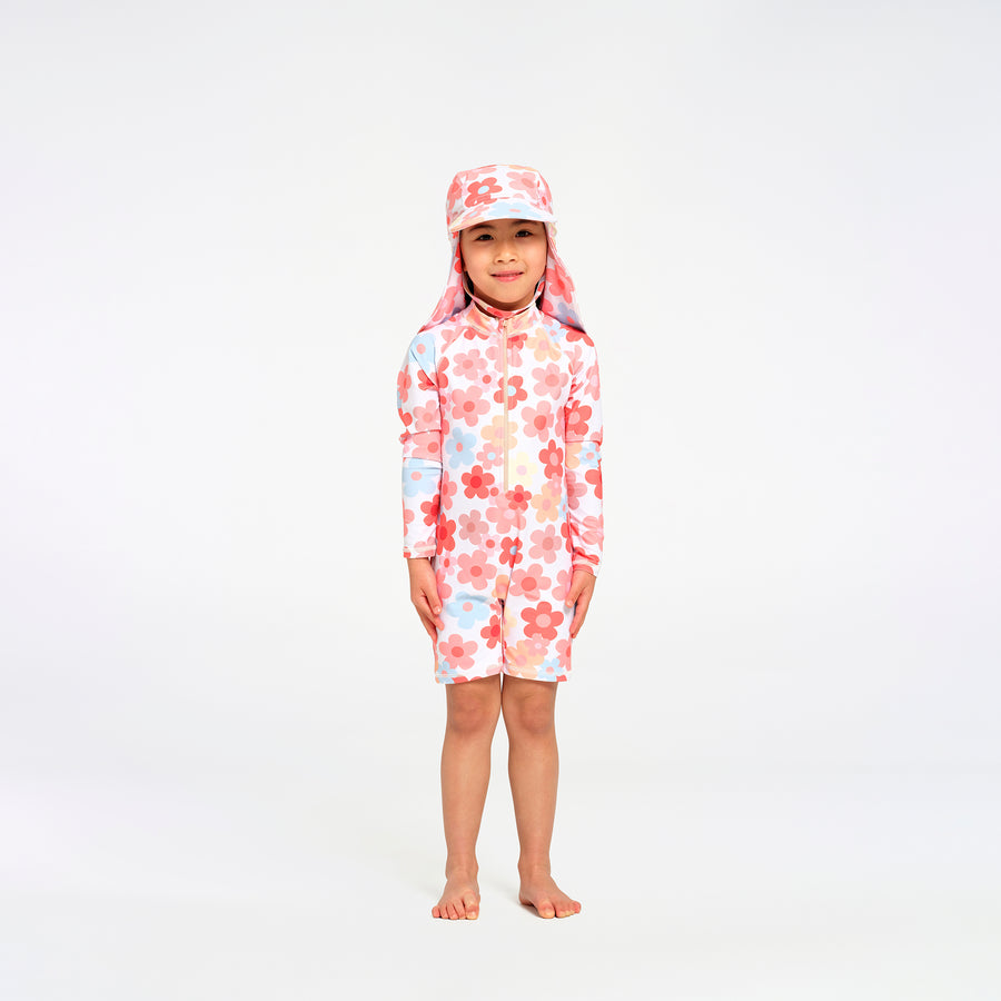 A young child standing barefoot against a plain white studio background, wearing a long-sleeve, short-legged swim suit with a matching sun hat. The swimwear has a white base with a pink and red floral pattern and small light blue accents.
