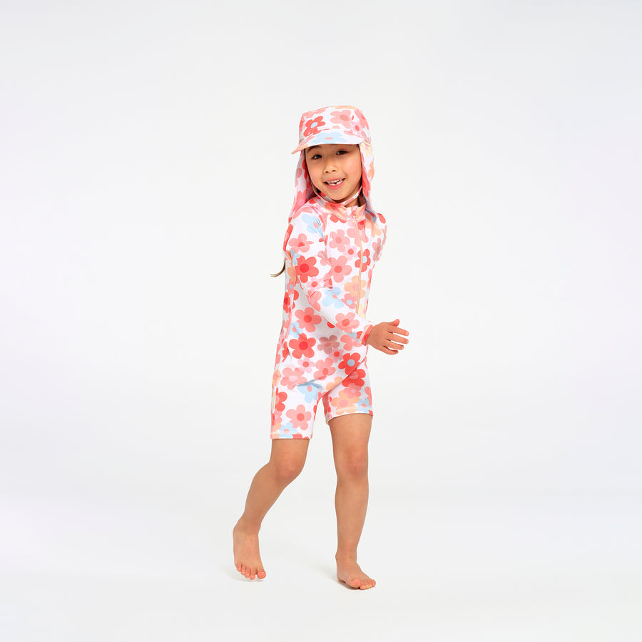 A young child wearing a one-piece floral swim suit with a matching sun hat, standing barefoot against a plain white studio background.