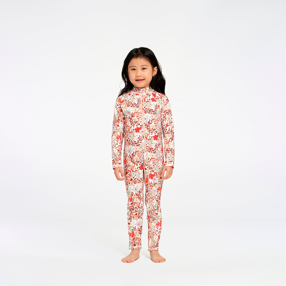 A young girl with long black hair stands barefoot against a plain white studio background, wearing a long-sleeve, full-length one-piece swimsuit with an allover floral print in red, orange, and white.