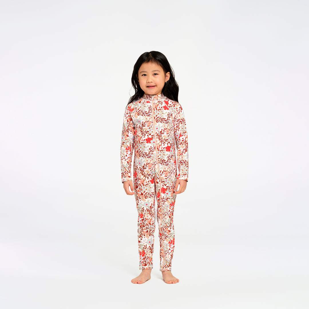 A young girl with long black hair stands barefoot against a plain white studio background, wearing a long-sleeve, full-length one-piece swimsuit with an allover floral print in red, orange, and white.