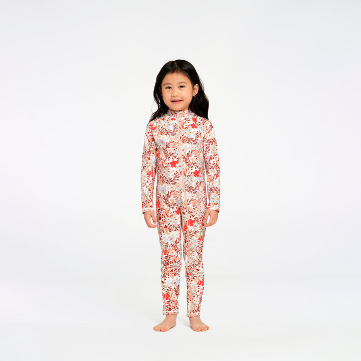 A young girl with long black hair stands barefoot against a plain white studio background, wearing a long-sleeve, full-length one-piece swimsuit with an allover floral print in red, orange, and white.