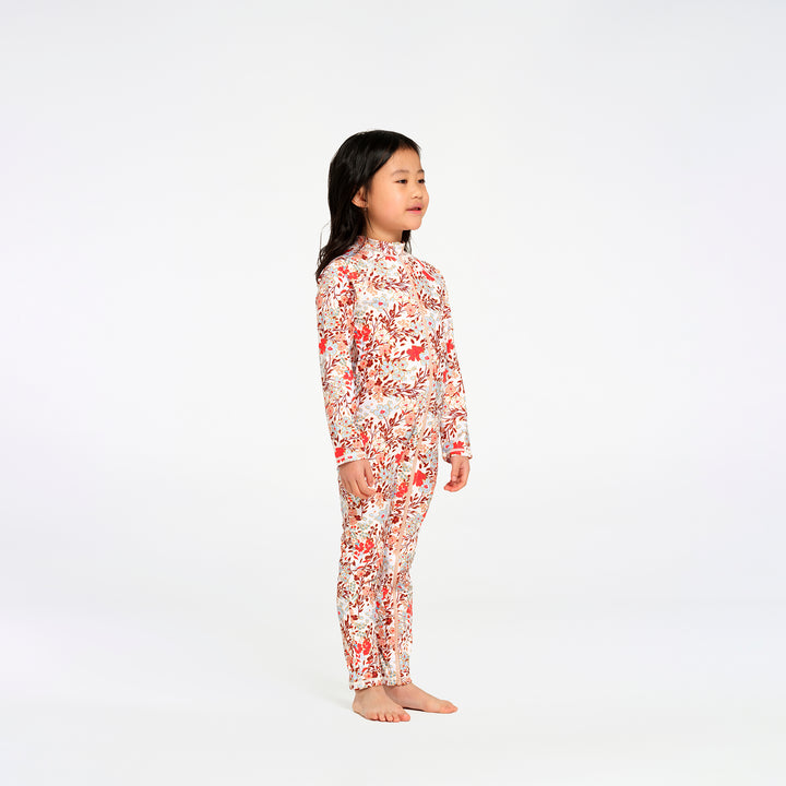 A young girl stands in profile wearing a long-sleeve, high-neck one-piece swimsuit with an all-over floral print in red, orange, and white on a light background, photographed against a seamless white studio backdrop.