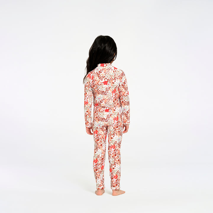 A child standing with their back to the camera, wearing a full body swimsuit with a colorful floral print set against a plain white studio backdrop.