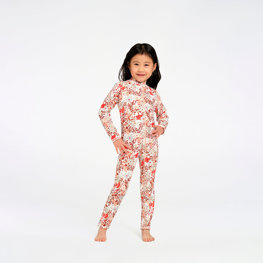 A young girl stands barefoot against a plain white studio background, wearing a long-sleeve, high-neck one-piece swimsuit with an all-over red and orange floral pattern on a white base.