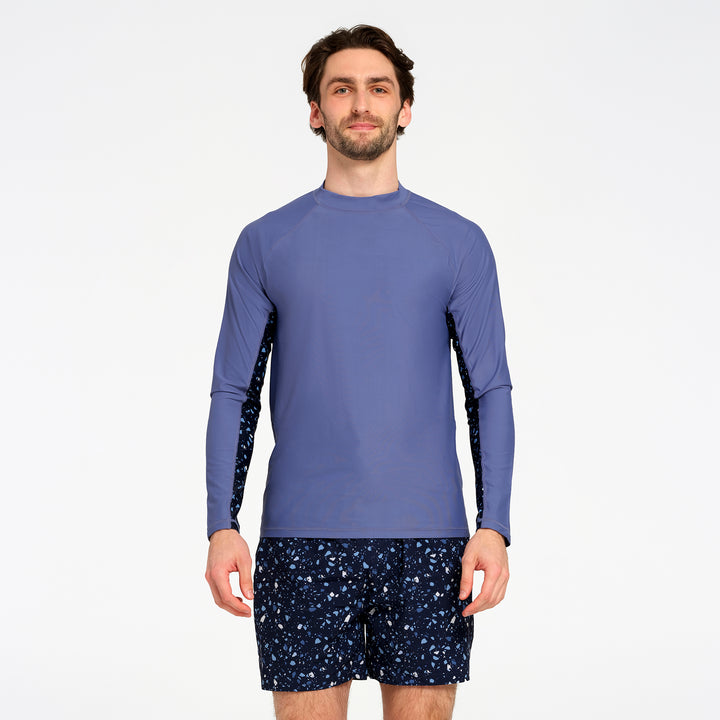 A man modeling a long-sleeve athletic rash guard in slate blue with dark patterned sleeves, paired with navy patterned swim shorts, against a plain white studio background.