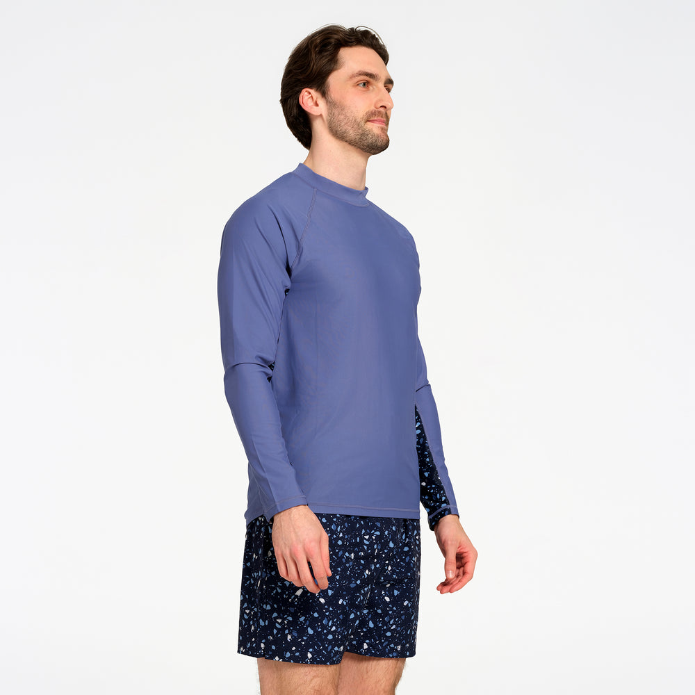 A man wearing a blue long-sleeve rash guard swim top and dark patterned swim shorts, shown from a side profile against a plain white studio background.