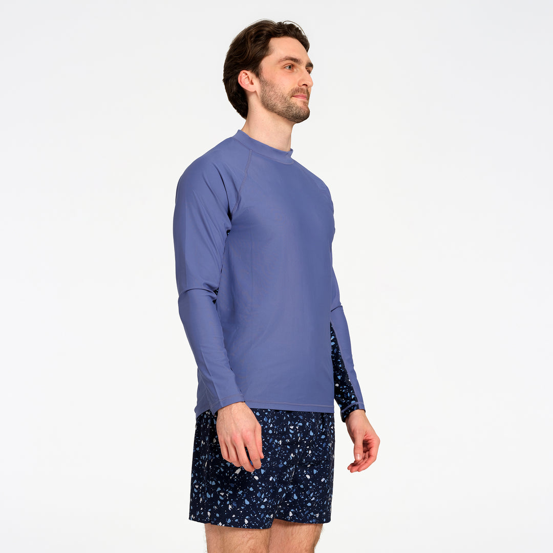 A man wearing a blue long-sleeve rash guard swim top and dark patterned swim shorts, shown from a side profile against a plain white studio background.