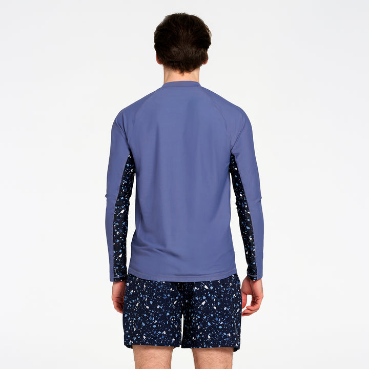 A man viewed from the back wearing a long-sleeve rash top in dusty blue with dark patterned sleeves and matching dark patterned shorts, against a plain white studio background.